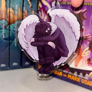 May include: A heart-shaped acrylic keychain featuring an illustration of a person with white angel wings, dark hair, and a purple outfit. The person is hunched over, with their head resting on their knees. The keychain has a black metal ring.