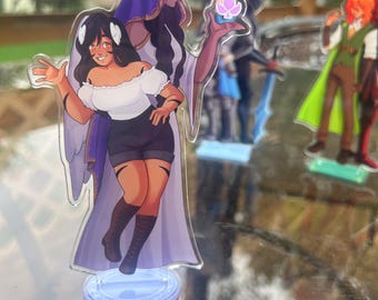 May include: A clear acrylic standee featuring a character with long dark hair, wearing a white off-shoulder top, black shorts, and dark boots, standing in front of a figure in a purple hooded cloak holding a glowing pink spade symbol icon.