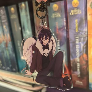 Op de afbeelding: Een heldere acryl sleutelhanger met een zittend anime-achtig personage met zwart haar, witte vleugels en een schedelontwerp op het shirt. Het personage draagt een bruine jas en een donkere broek. De sleutelhanger heeft een zwarte metalen sluiting.