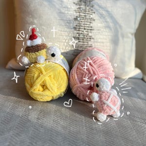 May include: A collection of handmade crochet items, including a cupcake with a Santa hat and two turtle figures. The items are placed on top of yarn balls in yellow and pink. A decorative pillow is in the background.