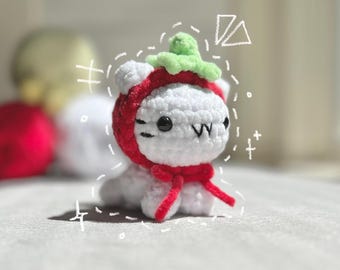 以下が含まれることがあります： 手作りの白いかぎ針編みの猫。赤いイチゴの帽子とスカーフを着用しています。猫は黒い目と縫い付けられた口をしています。イチゴの帽子には緑色の茎が付いています。猫は明るい灰色の表面に座っています。