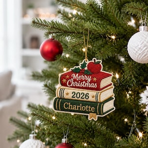 May include: A hanging Christmas tree ornament shaped like a stack of three books. The top red book reads Merry Christmas, the middle cream book reads 2026, and the bottom green book reads Charlotte. A red apple and green holly leaves sit on top.