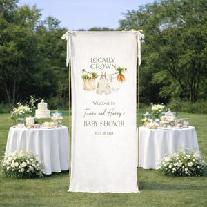 May include: A baby shower banner with the words "Locally Grown" and "Welcome to Tamara and Harry's Baby Shower" printed on it. The banner has illustrations of vegetables and baby clothes. The date of the baby shower is July 26, 2026.