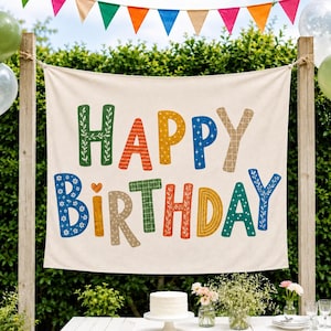 May include: A rectangular off-white fabric banner featuring the text HAPPY BIRTHDAY in large, multi-colored letters. Each letter displays a unique pattern, including floral motifs, stripes, dots, and stars in shades of green, orange, yellow, blue, and tan.