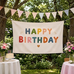 May include: A rectangular off-white fabric banner featuring the text HAPPY BIRTHDAY in multi-colored, patterned fabric letters with a small heart icon below. The banner is suspended outdoors with a string of triangular pennant flags above it.