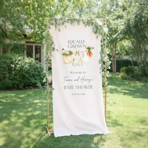 May include: A white fabric baby shower sign with the text "Locally Grown" and "Welcome to Tamara and Harry's Baby Shower, July 26, 2026." The sign is decorated with watercolor illustrations of vegetables and baby clothes. The sign is framed with a copper-colored metal structure and greenery.