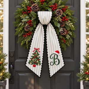 Peut inclure: Une couronne de fête composée d'aiguilles de pin vertes, de pommes de pin brunes et de baies rouges. Un long ruban en tissu rayé gris clair et blanc pend du centre, orné d'un sapin de Noël vert brodé et d'une grande lettre B avec un bonnet de Père Noël.