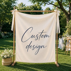 May include: A cream-colored fabric banner suspended from a wooden frame stand. The banner features the text "Custom design" in dark blue cursive script. The fabric is secured to the top horizontal wooden bar with white ribbons tied in bows.
