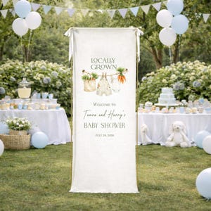 May include: A white banner with the text "Locally Grown" and "Welcome to Tamara and Harry's Baby Shower July 26, 2026." The banner has illustrations of vegetables and baby clothes. The background includes tables, balloons, and greenery.