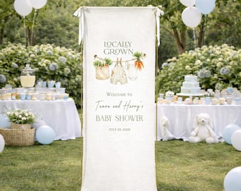 May include: A white banner with the text "Locally Grown" and "Welcome to Tamara and Harry's Baby Shower July 26, 2026." The banner has illustrations of vegetables and baby clothes. The background includes tables, balloons, and greenery.
