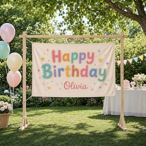 May include: A rectangular off-white fabric banner suspended on a wooden frame. The banner features the text "Happy Birthday" in colorful, rounded bubble letters, with the name "Olivia" printed below in a script font, surrounded by small stars.