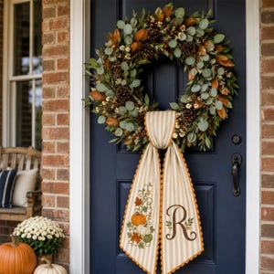 May include: A circular wreath featuring green eucalyptus leaves, brown magnolia leaves, and pinecones. A beige and white striped fabric banner hangs from the centre, decorated with embroidered pumpkins, autumn leaves, and the letter R in brown.