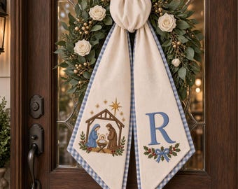 May include: A green wreath with white roses and gold berries features a cream-coloured fabric hanging with a blue gingham border. One side displays an embroidered nativity scene with a star, while the other shows a blue letter R above holly.