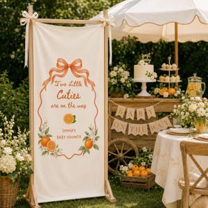 May include: A vertical cream-colored fabric banner suspended on a wooden frame, featuring an orange-colored bow and border, with the text "Two Little Cuties are on the way" and "EMMA'S BABY SHOWER" printed above and below orange fruit illustrations.
