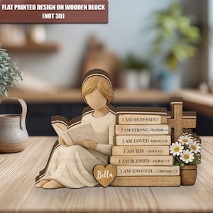May include: Wooden figurine of a woman reading, with stacked books and a cross. The books read "I AM REDEEMED", "I AM STRONG", "I AM LOVED", "I AM HIS", "I AM BLESSED", and "I AM ENOUGH". A heart with the name "Bella" is also present. The text "FLAT PRINTED DESIGN ON WOODEN BLOCK (NOT 3D)" is at the top.