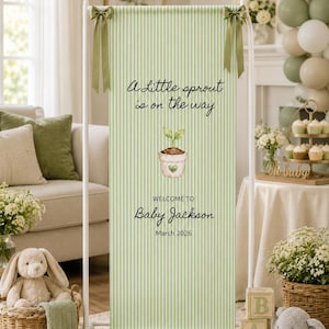 May include: A vertical rectangular fabric banner with a light green and white pinstripe pattern, suspended on a white metal stand with green ribbon bows. The text reads A Little sprout is on the way, WELCOME TO Baby Jackson, and March 2026.