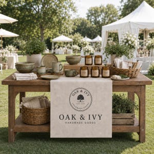 May include: A wooden display table featuring a beige runner with the text OAK & IVY HANDMADE GOODS and a tree logo. The table holds ceramic bowls, mugs, small glass jars, a woven basket with linens, and potted plants in an outdoor setting.