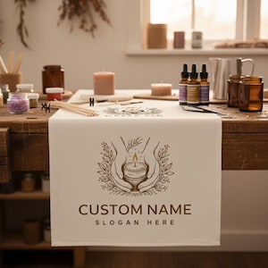 May include: Cream-colored table runner with a brown logo featuring hands holding a candle, with the text "CUSTOM NAME" and "SLOGAN HERE". The runner is on a wooden table with candles, jars, and essential oil bottles.