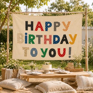 May include: A rectangular off-white fabric banner featuring the text HAPPY BIRTHDAY TO YOU! in large, multi-colored letters with various patterns including polka dots, stripes, stars, and gingham checks, suspended on a wooden frame.