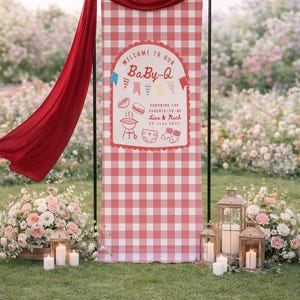 May include: A red and white gingham banner with the text "WELCOME TO OUR BaBy-Q" is displayed at an outdoor event. The banner includes illustrations of a grill, hot dog, burger, diaper, and socks. A red fabric drapes from the top. Lanterns and flowers are in the foreground.