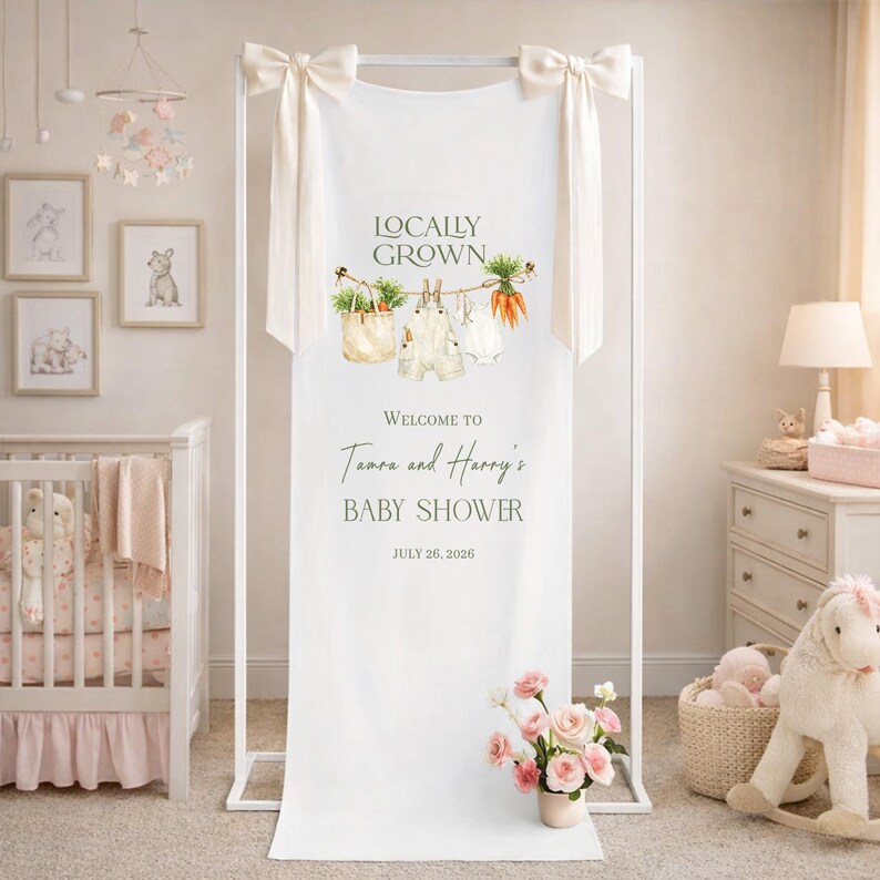 May include: A white fabric baby shower banner with the text "Locally Grown" and "Welcome to Tamara and Harry's Baby Shower July 26, 2026." The banner is decorated with illustrations of vegetables and baby clothes.