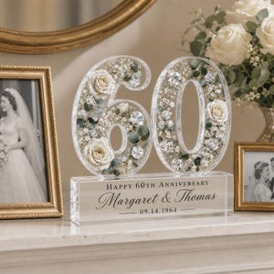 May include: A clear acrylic number 60 filled with preserved white roses, greenery, and small white flowers. The base is a rectangular block with the text "HAPPY 60TH ANNIVERSARY", "Margaret & Thomas", and "09.14.1964" printed in black.