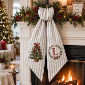 Peut inclure: Une écharpe de cheminée en tissu rayé gris et blanc avec deux pans suspendus. Un pan présente un sapin de Noël brodé avec un nœud rouge, tandis que l'autre arbore une lettre L rouge dans une couronne verte avec un petit nœud rouge à la base.