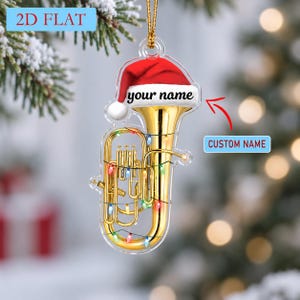Peut inclure: Une décoration de Noël en acrylique transparent plat en forme de tuba doré. Le tuba est entouré de guirlandes lumineuses colorées et surmonté d'un bonnet de Père Noël rouge et blanc. Le bonnet porte le texte your name en noir.