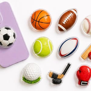 May include: A collection of sports-themed phone grips, including a basketball, football, baseball, tennis ball, rugby ball, cricket bat and ball, golf ball, hockey stick and puck, and a boxing glove. A purple phone with a soccer ball grip is also present.