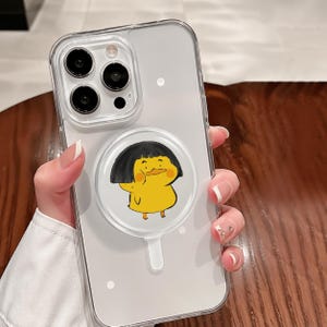 May include: Clear phone case with a cartoon duck design. The case is transparent, showcasing the phone's features. The duck is yellow with black hair and a cute expression. The phone case is held by a person.