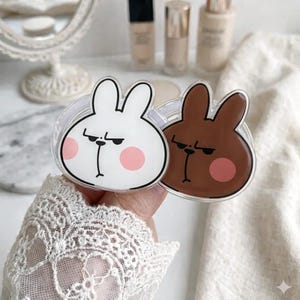 May include: Two clear phone grips featuring cartoon bunny faces. One is white with a black outline and pink cheeks, the other is brown with a black outline and pink cheeks. The grips have a clear, circular base.