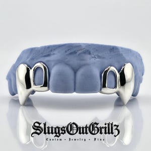 May include: A set of silver-colored vampire fang grillz fitted onto a light blue dental mold. The metal teeth covers feature pointed canine designs. Below the dental mold, the black text reads SlugsOutGrillz Custom Jewelry Fine.