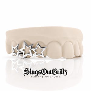 May include: A silver-toned dental grill featuring three open-star designs attached to a beige dental mold. The text SlugsOutGrillz Custom Jewelry Fine is printed in black below the mold. The grill covers three teeth on the dental model.