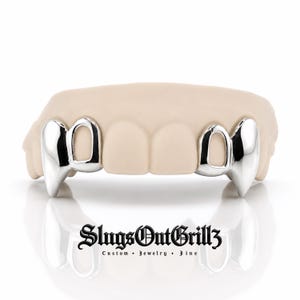 May include: A set of silver-colored vampire fang grillz fitted over two teeth on each side of a beige dental mold. The text SlugsOutGrillz Custom Jewelry Fine is printed in black in a gothic-style font below the dental appliance.