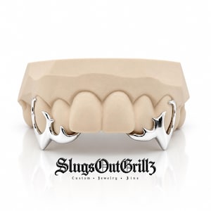 May include: A set of silver-colored metal tooth covers shaped like sharp fangs attached to a beige dental mold. The text SlugsOutGrillz Custom Jewelry Fine is printed in black below the mold. The design features pointed edges and a smooth finish.