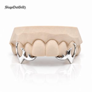 May include: A set of silver-colored metal teeth grills featuring a sharp, pointed design on the canine teeth. The text SlugsOutGrillz is visible in the upper left corner. The grills are fitted onto a beige dental mold of human teeth.