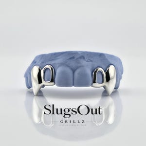 May include: Silver-colored fang grillz, displayed on a blue dental mold. The grillz have a polished finish and a pointed, vampire-like design. The brand name "SlugsOut GRILLZ" is visible below the grillz.