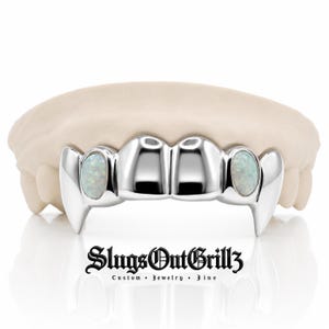May include: A silver-colored dental grill featuring four teeth with pointed fang-like ends on the outer edges. Two oval-shaped iridescent white opals are set into the teeth adjacent to the fangs. The text SlugsOutGrillz Custom Jewelry Fine is below.