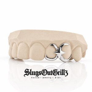 May include: A silver-colored tooth grill featuring a central four-pointed star design flanked by two crescent moon shapes, fitted onto a beige dental mold. Below the mold, the text reads SlugsOutGrillz Custom Jewelry Fine in black lettering.