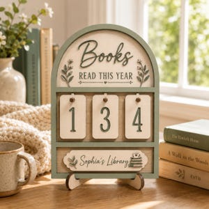May include: An arched wooden book tracker sign with a sage green frame and cream background. It features the text "Books READ THIS YEAR" above three hanging number tags displaying "134" and a bottom plaque reading "Sophia's Library" with a book icon.