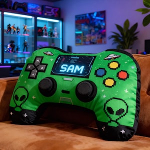 May include: A green, plush video game controller-shaped pillow with black accents. The pillow features a digital display with the name "SAM" and pixelated alien graphics. The controller has black joysticks and directional buttons.