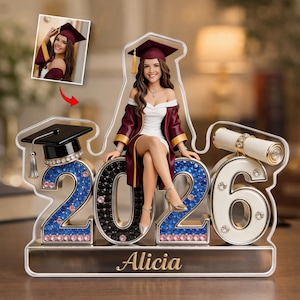 May include: A graduation-themed figurine featuring a young woman in a maroon graduation gown and cap, seated on the numbers "2026". The numbers are adorned with rhinestones. A small photo is included. The name "Alicia" is displayed on the base.