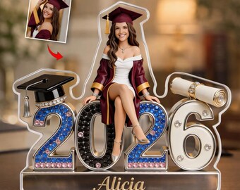 May include: A graduation-themed figurine featuring a young woman in a maroon graduation gown and cap, seated on the numbers "2026". The numbers are adorned with rhinestones. A small photo is included. The name "Alicia" is displayed on the base.