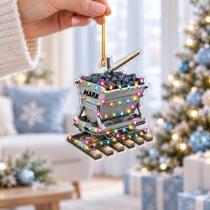 May include: A flat, clear acrylic Christmas ornament shaped like a mining cart filled with black coal, featuring the name MARK printed on the side. The cart is decorated with colorful string lights and sits on wooden tracks.