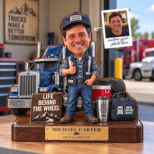 May include: A custom bobblehead figurine of a truck driver wearing a blue cap, plaid shirt, and vest, standing before a blue semi-truck model. The base features a plaque reading MICHAEL CARTER TRUCK DRIVER and a sign saying LIFE BEHIND THE WHEEL.