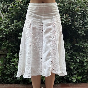 May include: A white midi skirt featuring vertical panels of intricate crochet lace inserts alternating with solid fabric sections. The solid panels are decorated with delicate white floral embroidery, and the skirt has a high-waisted fit.