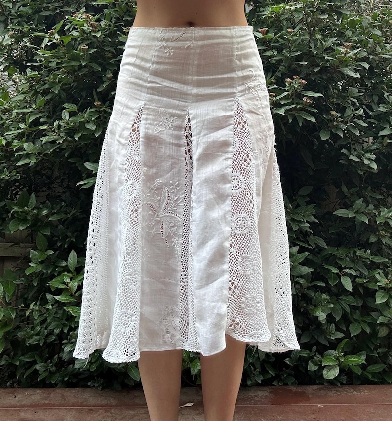 May include: A white midi skirt featuring vertical panels of intricate crochet lace inserts alternating with solid fabric sections. The solid panels are decorated with delicate white floral embroidery, and the skirt has a high-waisted fit.