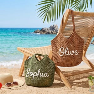May include: A beach scene with a woven chair, two crochet tote bags, a straw hat, and a cocktail. One bag is brown with "Olive" in white, the other is green with "Sophia". The hat has sunglasses. A red hibiscus flower is on the sand.