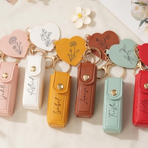 Può includere: Una collezione di portachiavi personalizzati con targhette a forma di cuore con disegni floreali e custodie in similpelle abbinate. Le custodie sono in vari colori, ognuna con un nome stampato. I portachiavi hanno finiture color oro.