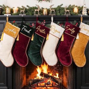 Könnte beinhalten: Sechs Weihnachtssocken aus Samt hängen in einer Reihe, mit den auf die Bündchen gestickten Namen Sophia, Frank, Austin, Regan, Emily und Darcy. Die Socken sind in Senfgelb, Dunkelgrün und Dunkelrot gehalten und haben weiße Akzente.